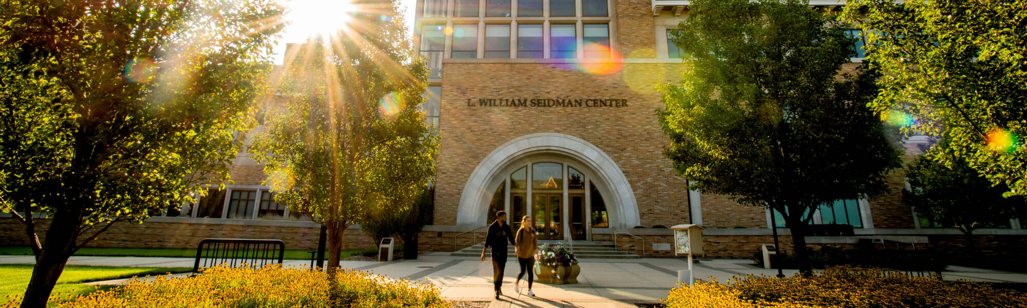 Two students walking in front of the Seidman Center building with the morning sun rising behind the trees